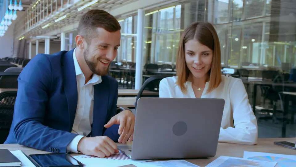 Duas pessoas colegas de trabalho revisam juntas um relatório no notebook, sorrindo